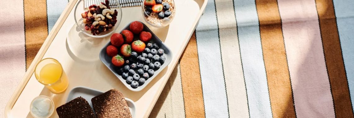 Top view of a healthy breakfast tray with fruits, bread, and juice on a striped tablecloth.